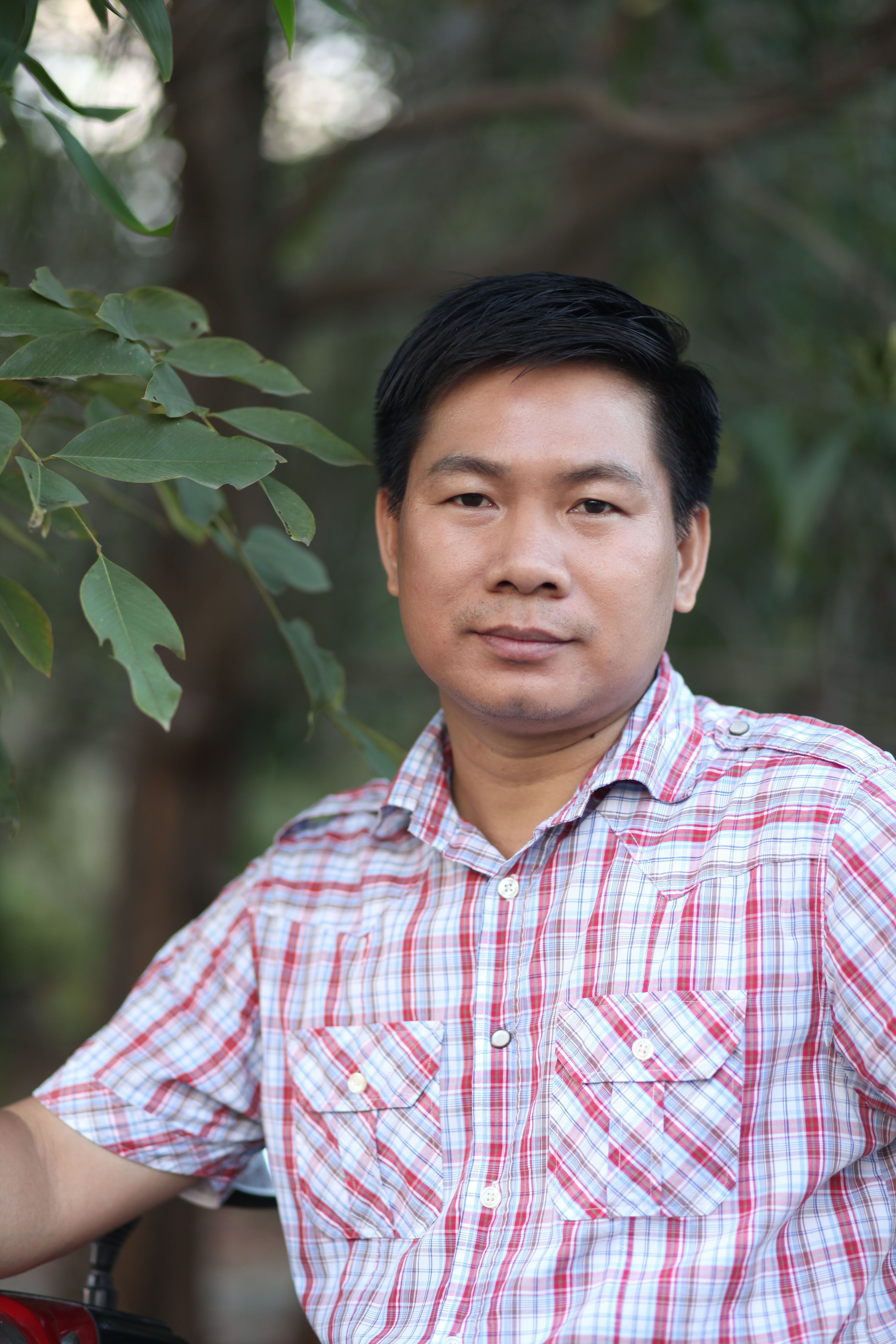 Man in a plaid shirt posing calmly outdoors with greenery.