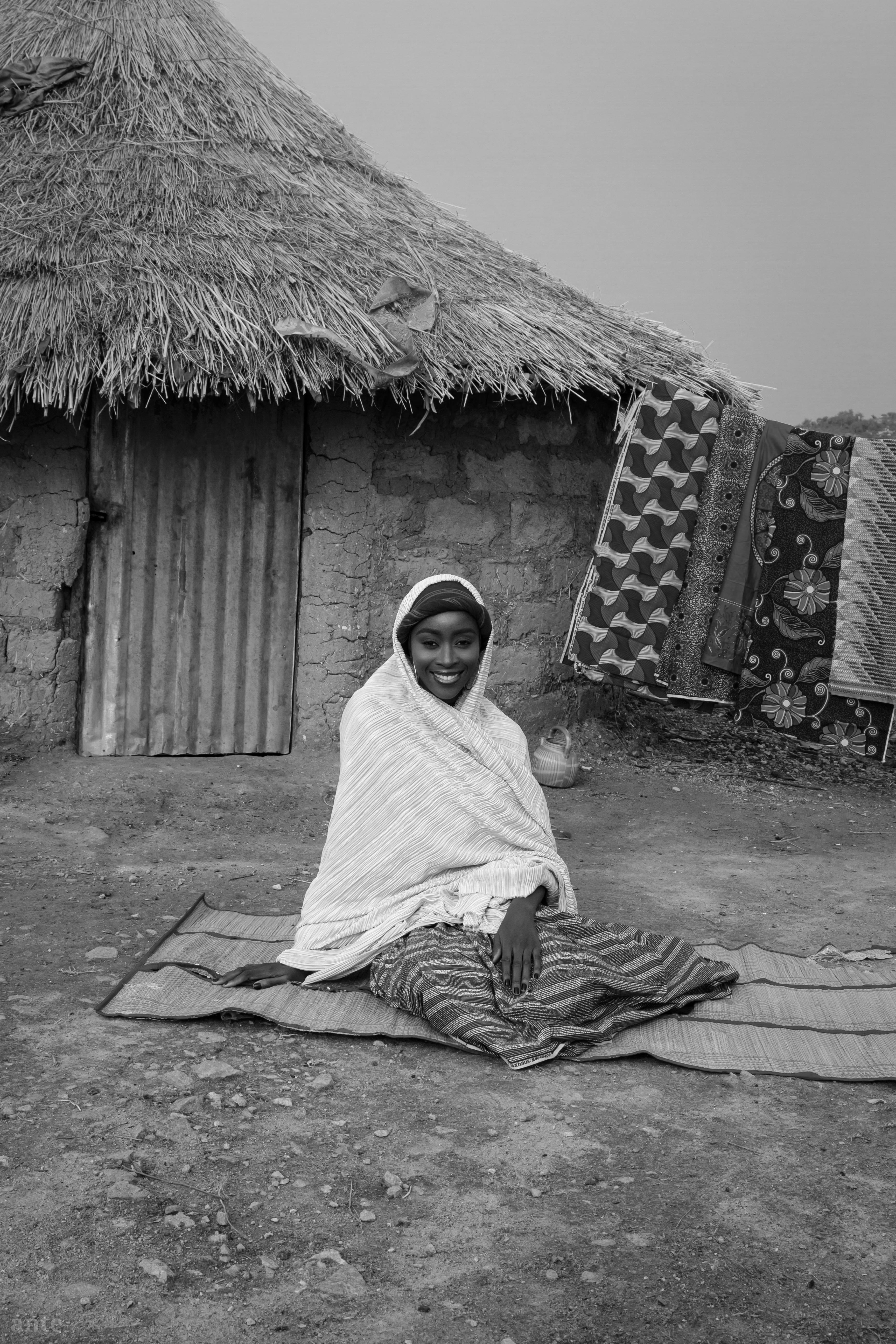Black and white portrait of a woman seated outside a rural hut, smiling warmly.