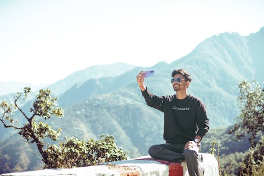 A young man captures a selfie with scenic mountains in the background on a sunny day.