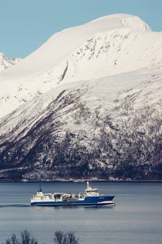 A fishing vessel glides through a tranquil Norwegian fjord surrounded by snowy mountains.