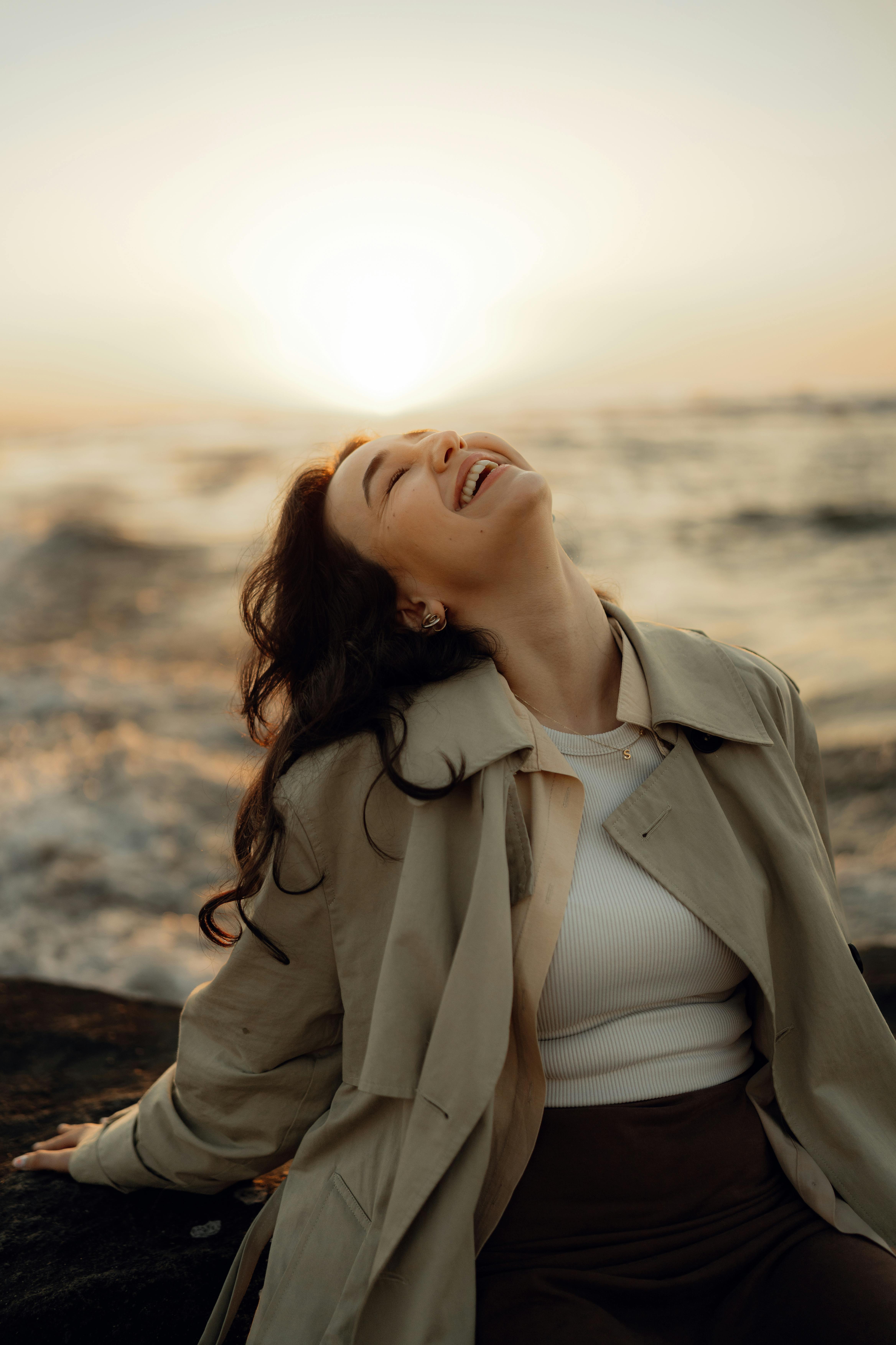 Gratis Una mujer con gabardina disfruta de una puesta de sol junto al océano, exudando alegría y serenidad. Foto de stock