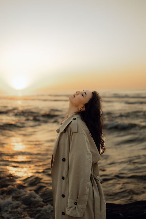 Free A woman in a beige coat enjoys the serene ocean sunset, expressing freedom and peace. Stock Photo