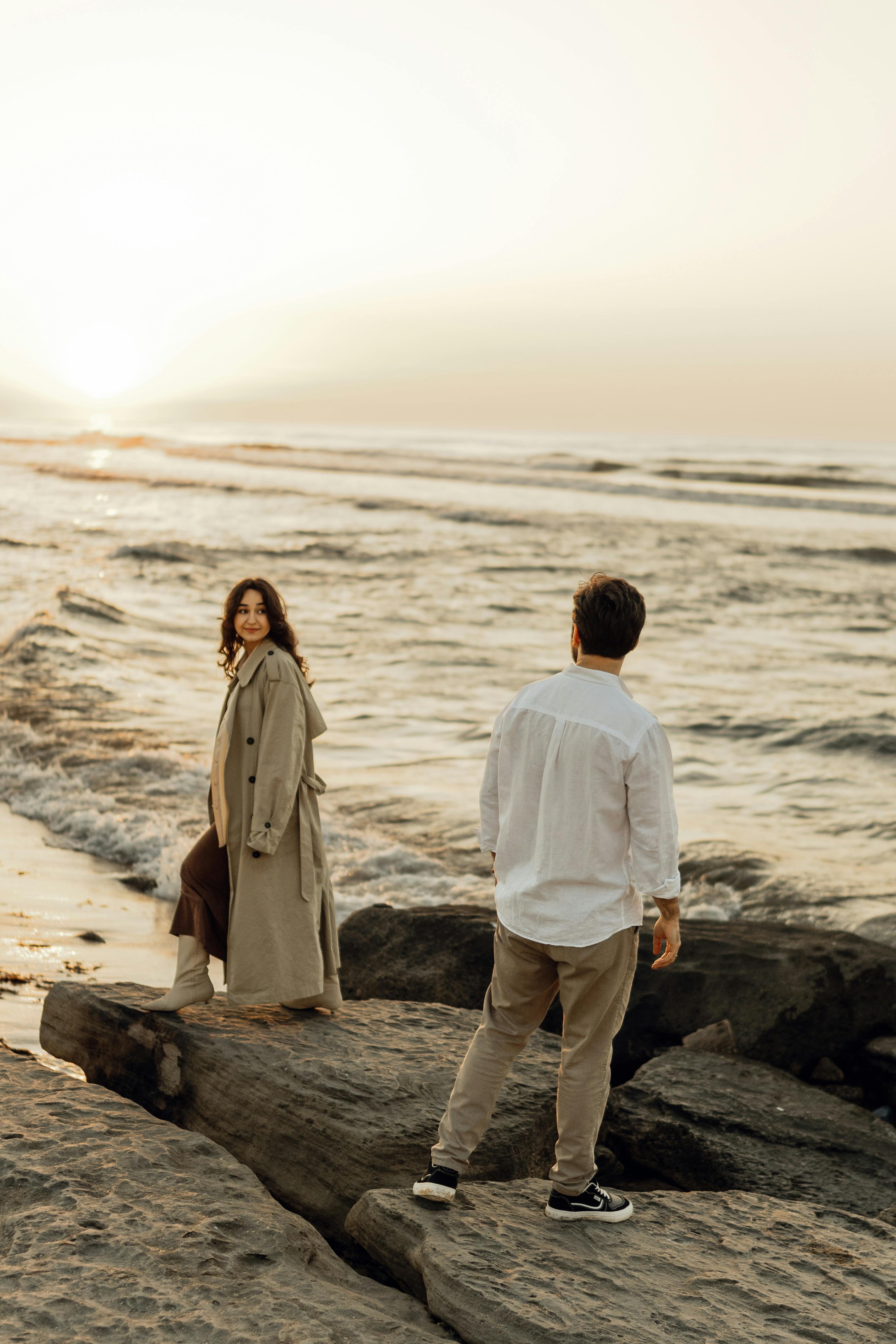 A couple walks along rocky shores with a beautiful ocean sunset backdrop, evoking romance and tranquility.