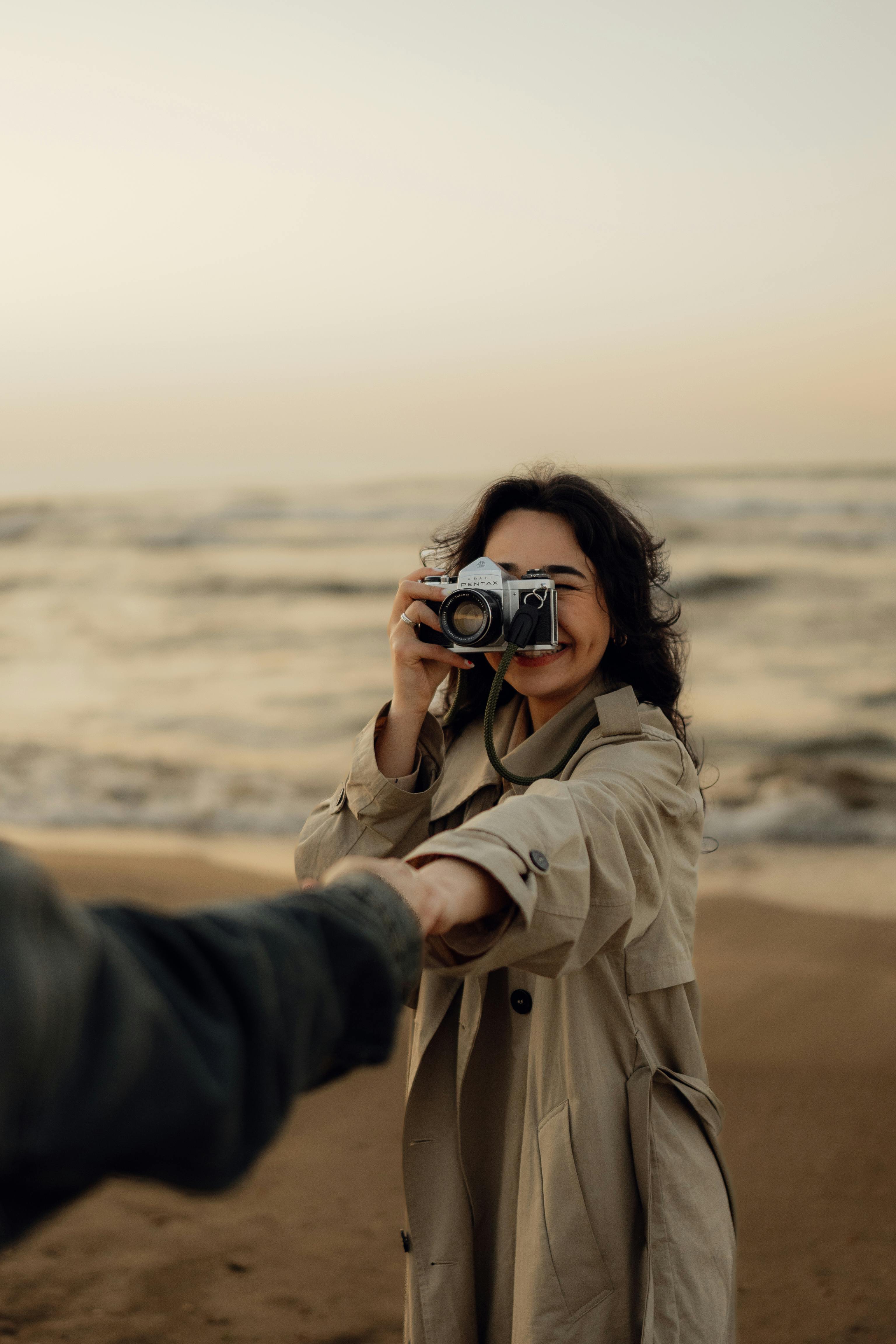 A woman in a trench coat holds a camera on a breezy beach, capturing life's moments at sunset.