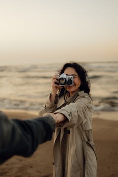 A woman in a trench coat holds a camera on a breezy beach, capturing life's moments at sunset.