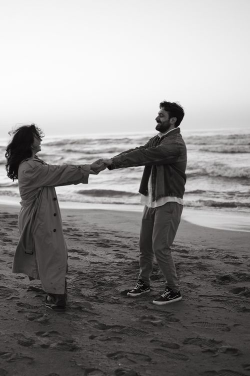 Free A couple dances happily on a beach shoreline in black and white, capturing a serene moment of joy at sunrise. Stock Photo