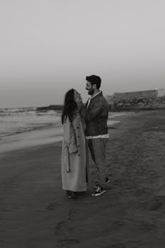 A couple enjoys a romantic moment on the beach at twilight, captured in a moody black and white setting.