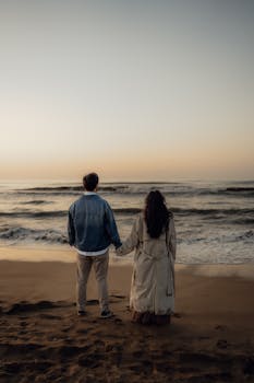 Serene scene of a couple holding hands while facing the ocean at sunset.