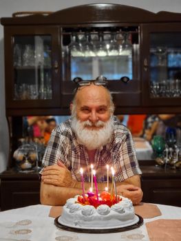 Senior man enjoys his birthday with candles lit on cake, surrounded by family indoors.