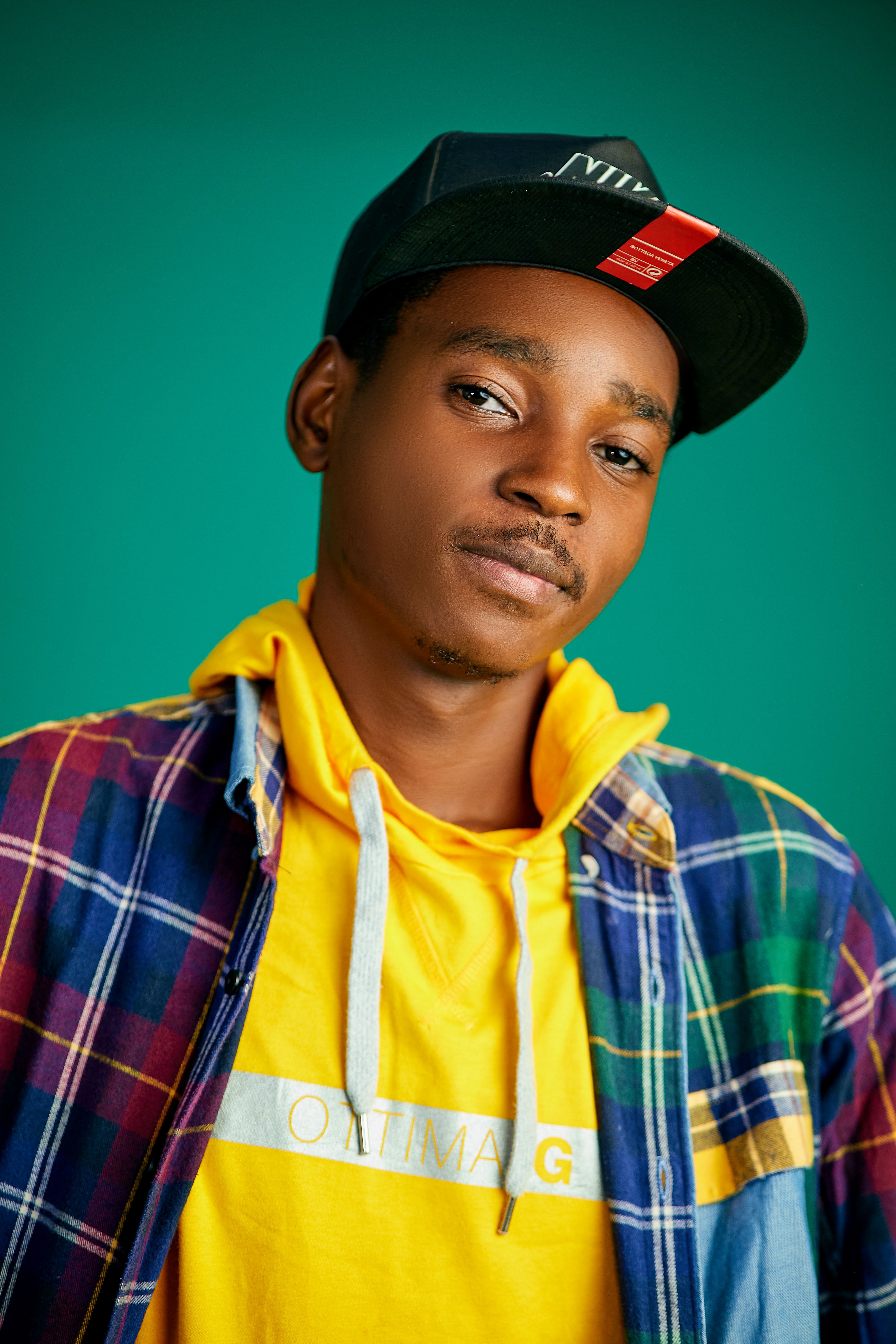 Portrait of a stylish young adult in a plaid jacket and cap against a green background.