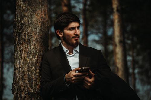 Free Businessman in nature, holding a phone, leaning against a tree. Stock Photo