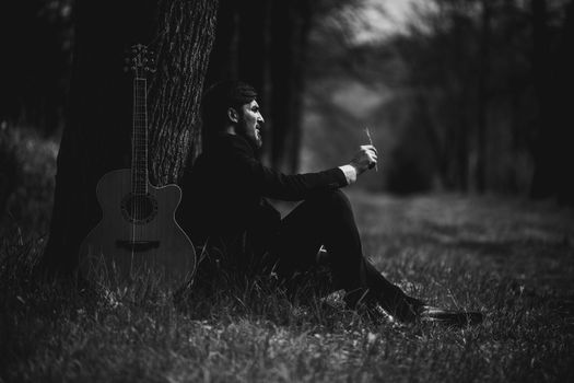 Black and white image of man with guitar under tree. Contemplative atmosphere.
