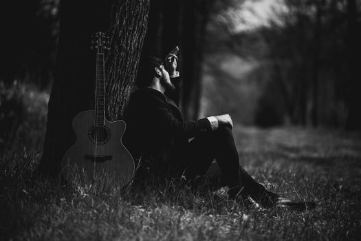 A contemplative man with a guitar sits under a tree in a forest.