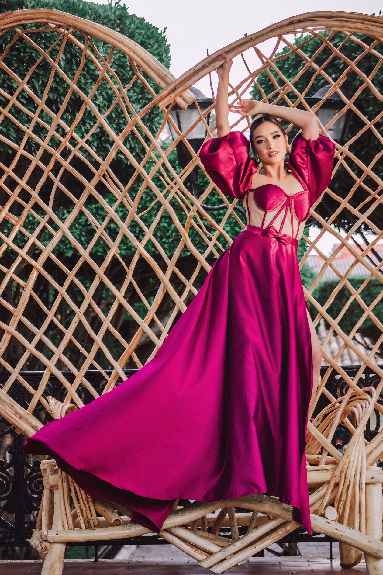 Woman in a magenta satin gown with puff sleeves and corset lacing, posing on a woven bench