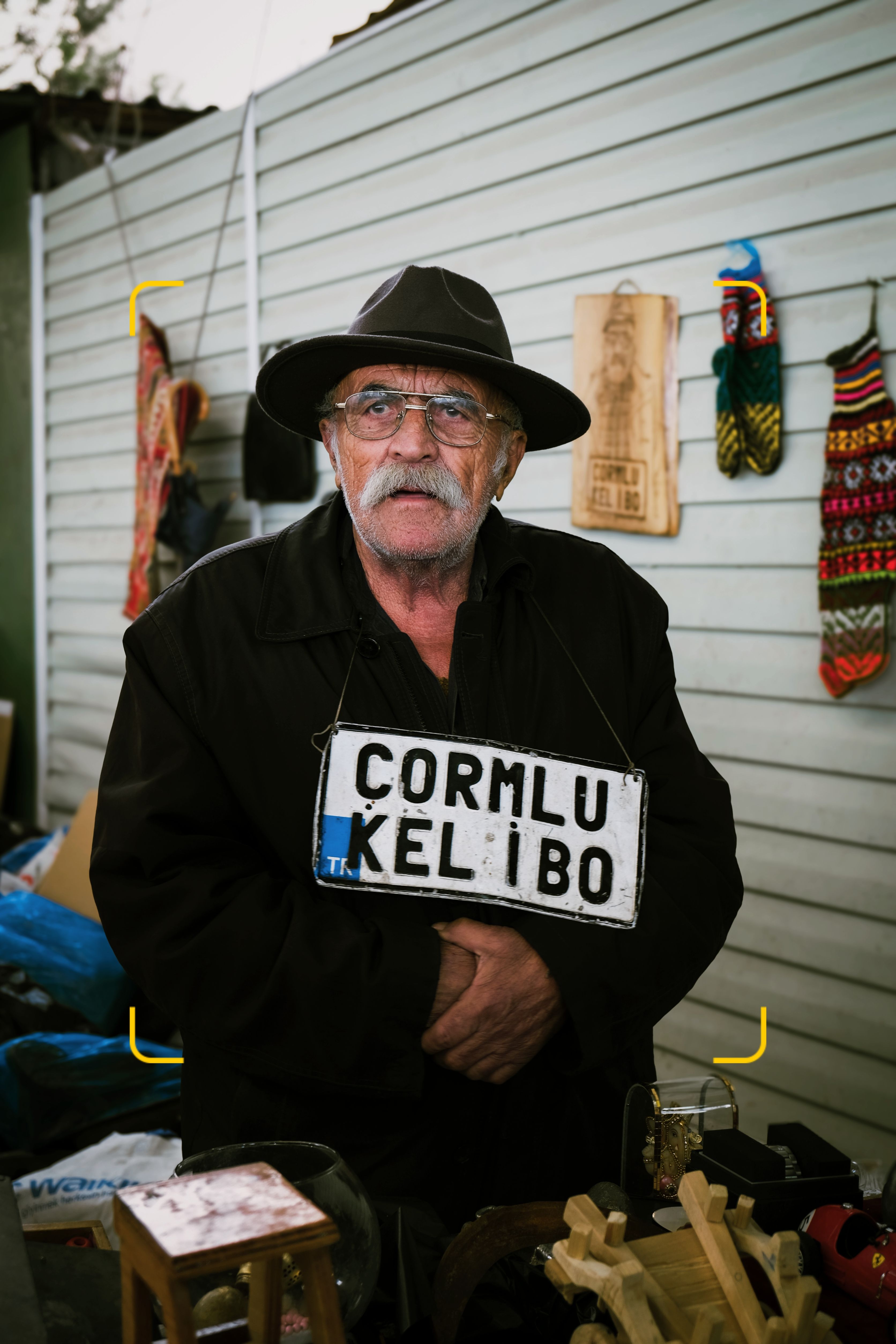Elderly man in a coat and hat holding a vintage sign at an outdoor market.