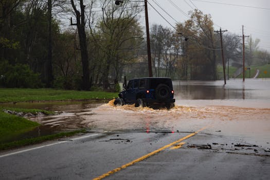 A jeep traverses a flooded road surrounded by trees, showcasing a challenging weather condition.