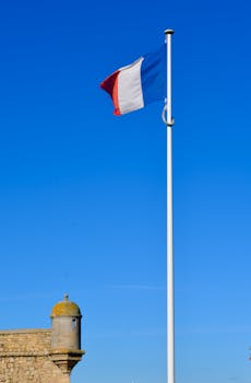 A French flag waving on a tall flagpole beside a historic stone structure under a clear blue sky.