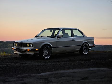 Vintage BMW E30 parked at sunset with scenic background, ideal for automotive enthusiasts.