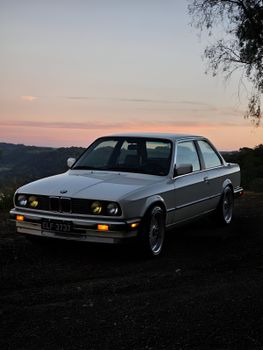 A white classic car parked outdoors with mountains and a sunset in the background.
