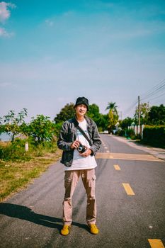 A young man stands on a road with trees, holding a camera on a sunny day, posing confidently.