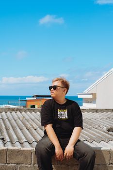 A young man sits on a rooftop looking at the ocean, wearing sunglasses under a clear blue sky.