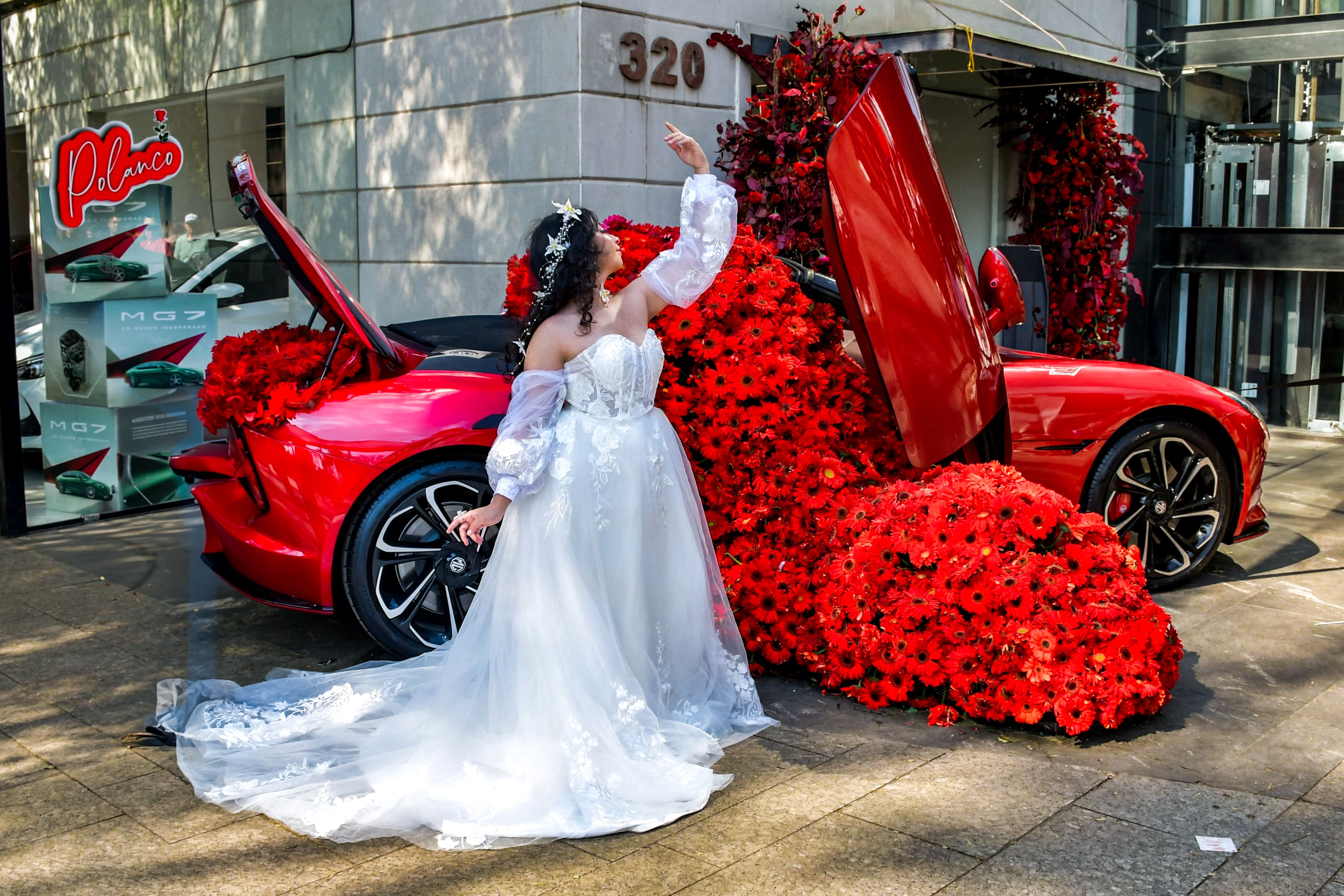 A bride in a white gown poses with a red sports car adorned with flowers, creating a stunning visual.