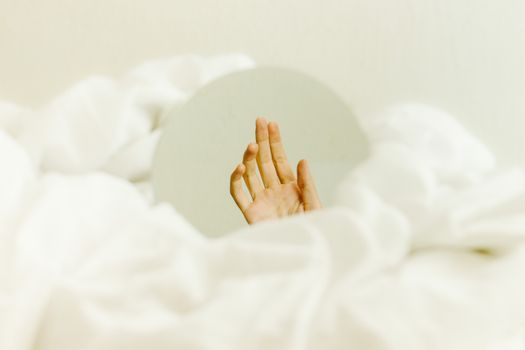 A hand reflected in a mirror surrounded by white sheets, creating a serene minimalist scene.