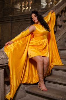Stylish woman poses in a flowing yellow dress on ornate stone stairs, exuding elegance.