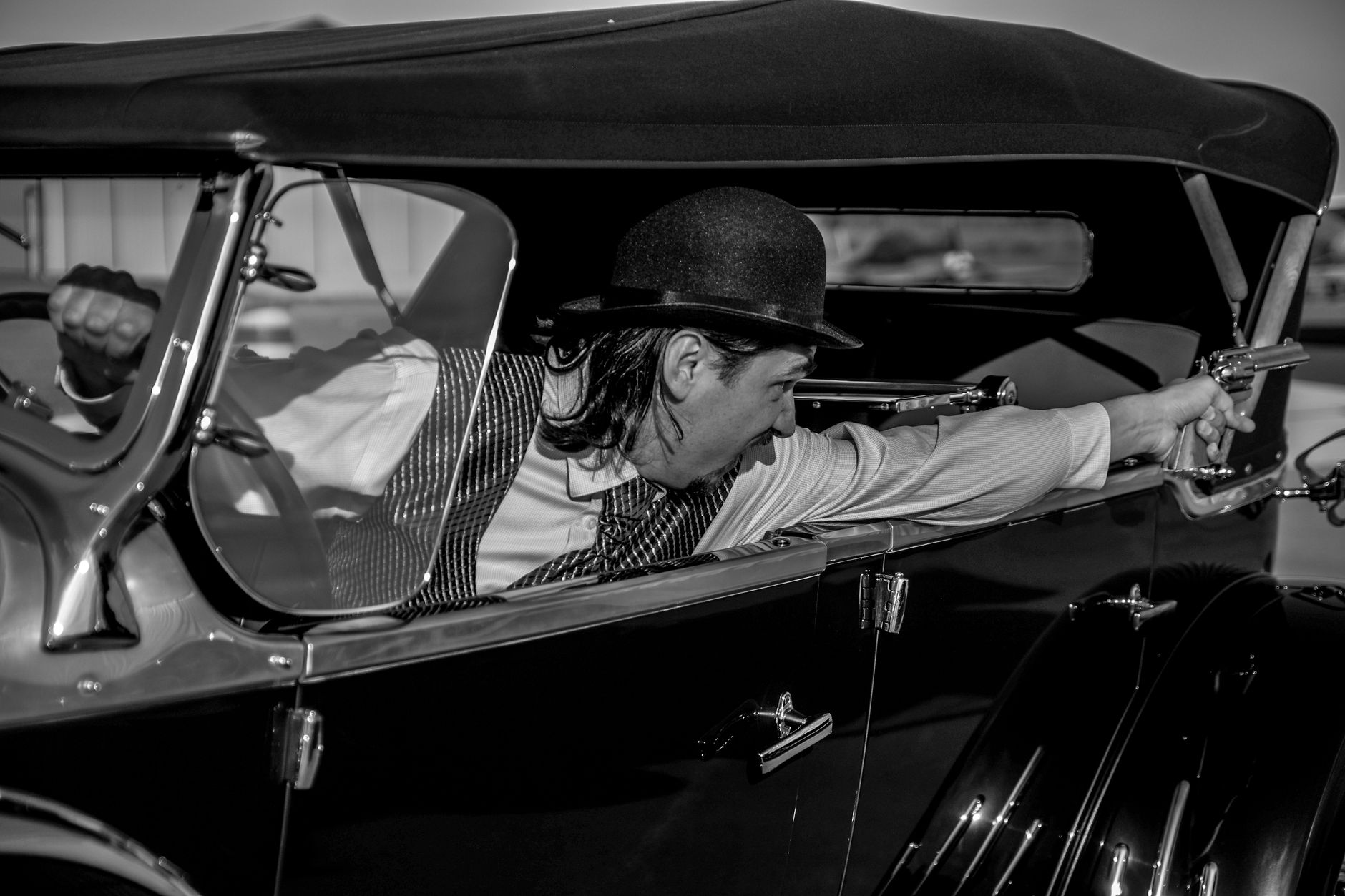 Dramatic black and white photo of a vintage 1930s-style gangster aiming a pistol from a classic car.
