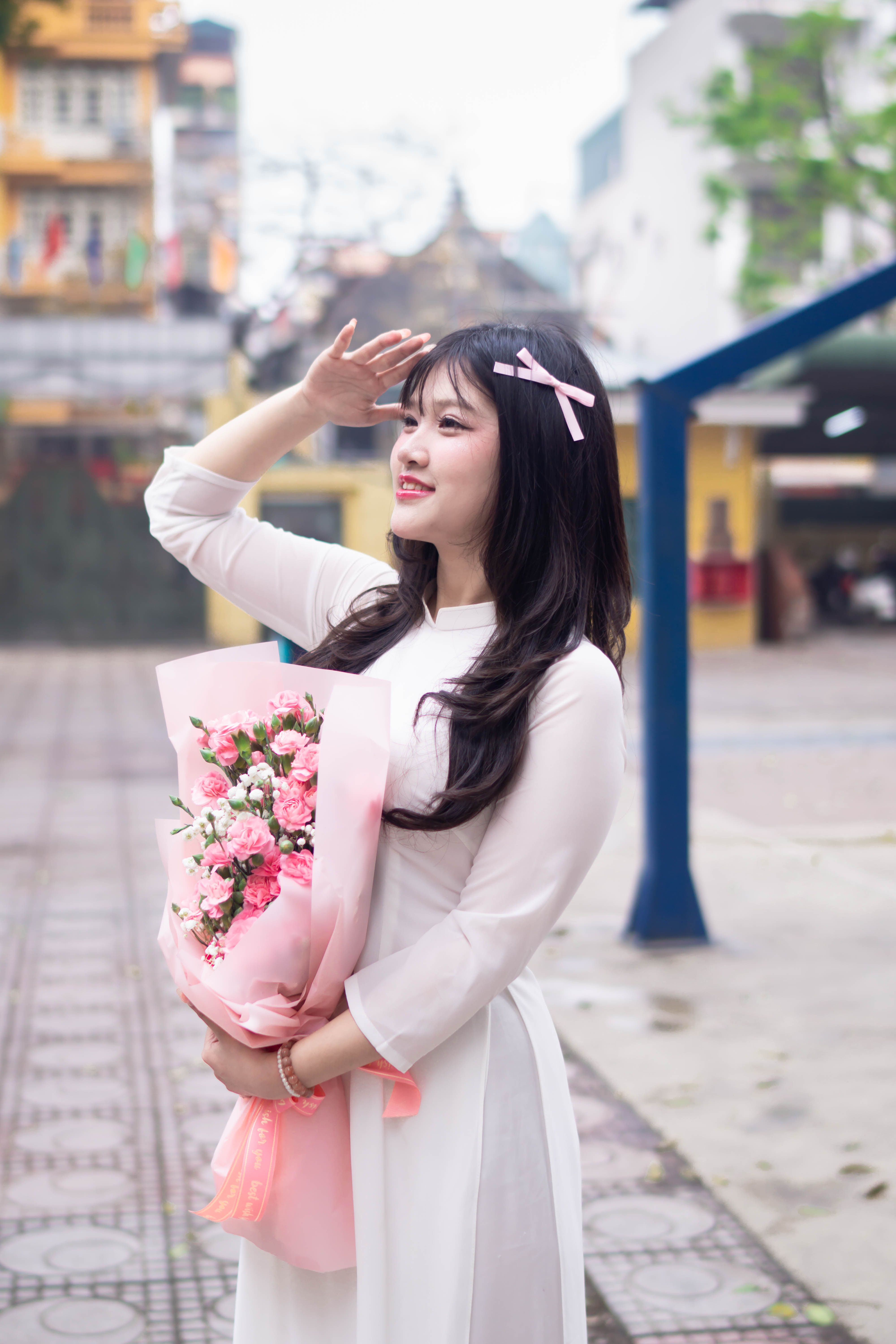 Young woman in traditional dress holds a beautiful pink bouquet, smiling in an outdoor city setting.