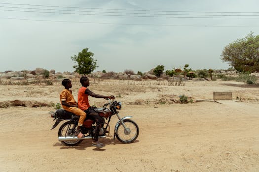 Two individuals riding a motorcycle on a dusty road in a rural landscape during day.