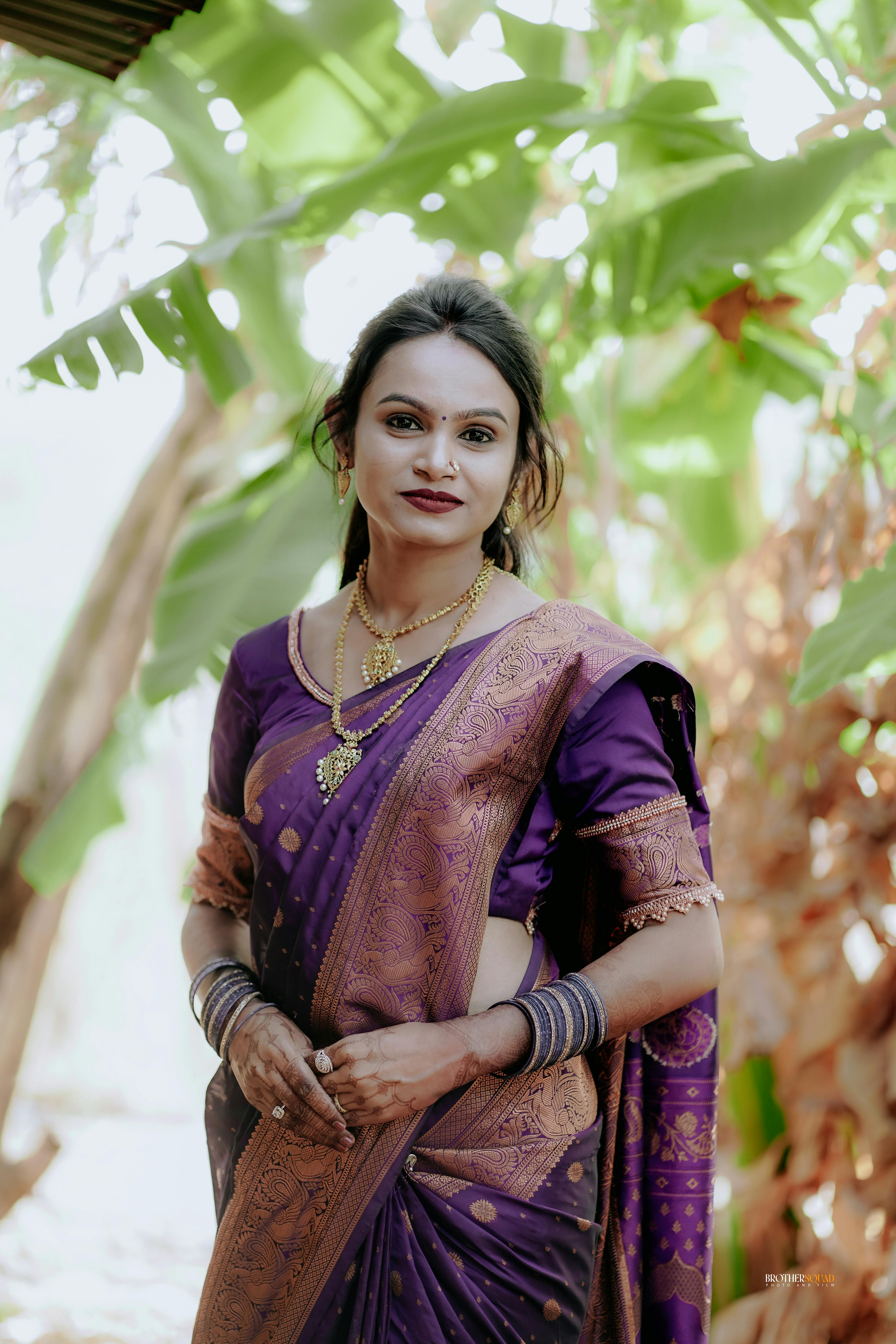 A woman in a purple sari poses gracefully outdoors, surrounded by lush greenery.
