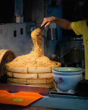 A street vendor serving a large bamboo steamer filled with Asian noodles in a bustling market.
