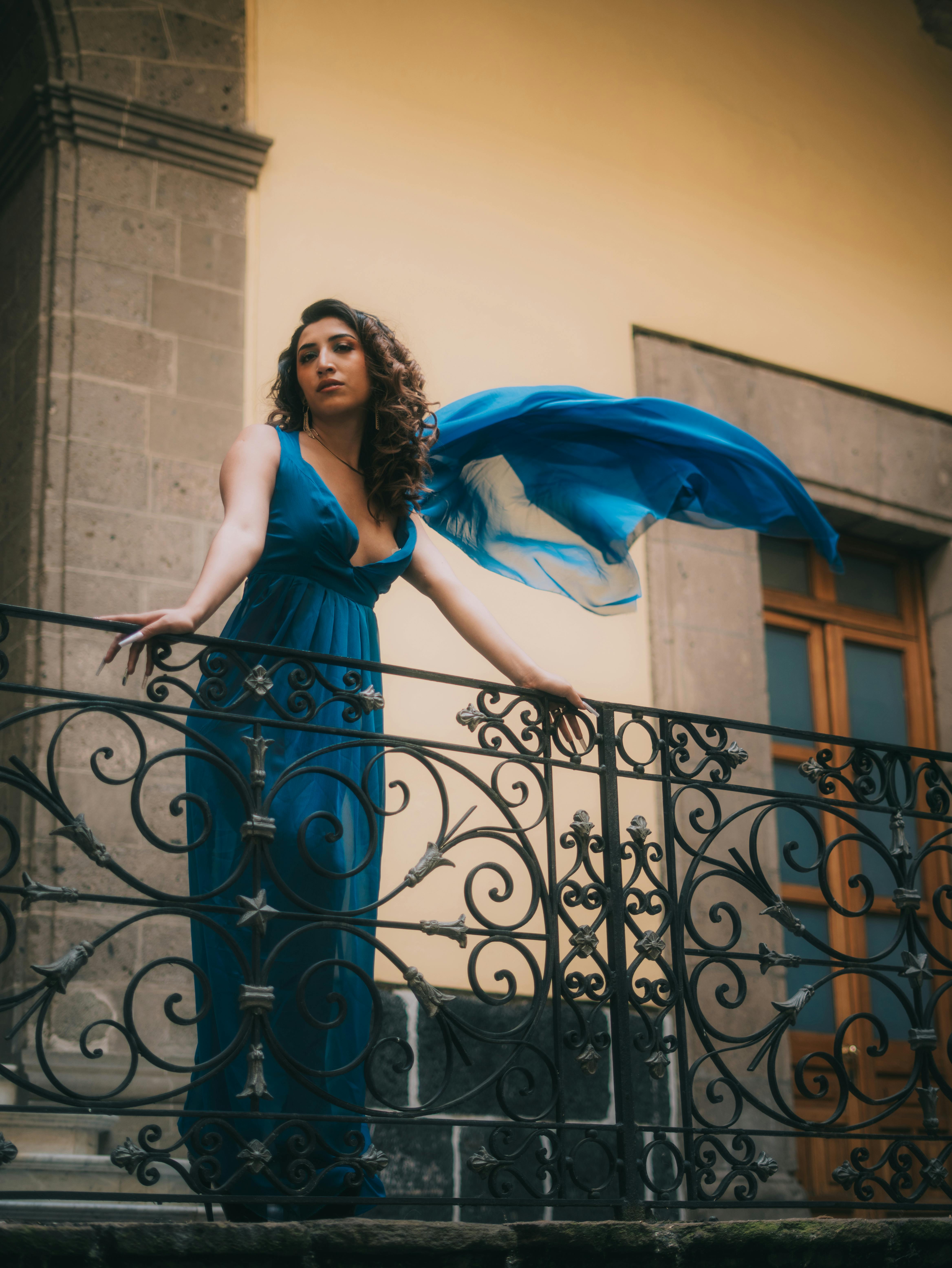 De franc Una dona equilibrada amb un vestit blau es troba en un balcó històric amb teixit fluid. Foto d'estoc