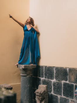 A fashion model in a blue dress poses dramatically on a stone pillar inside a museum.