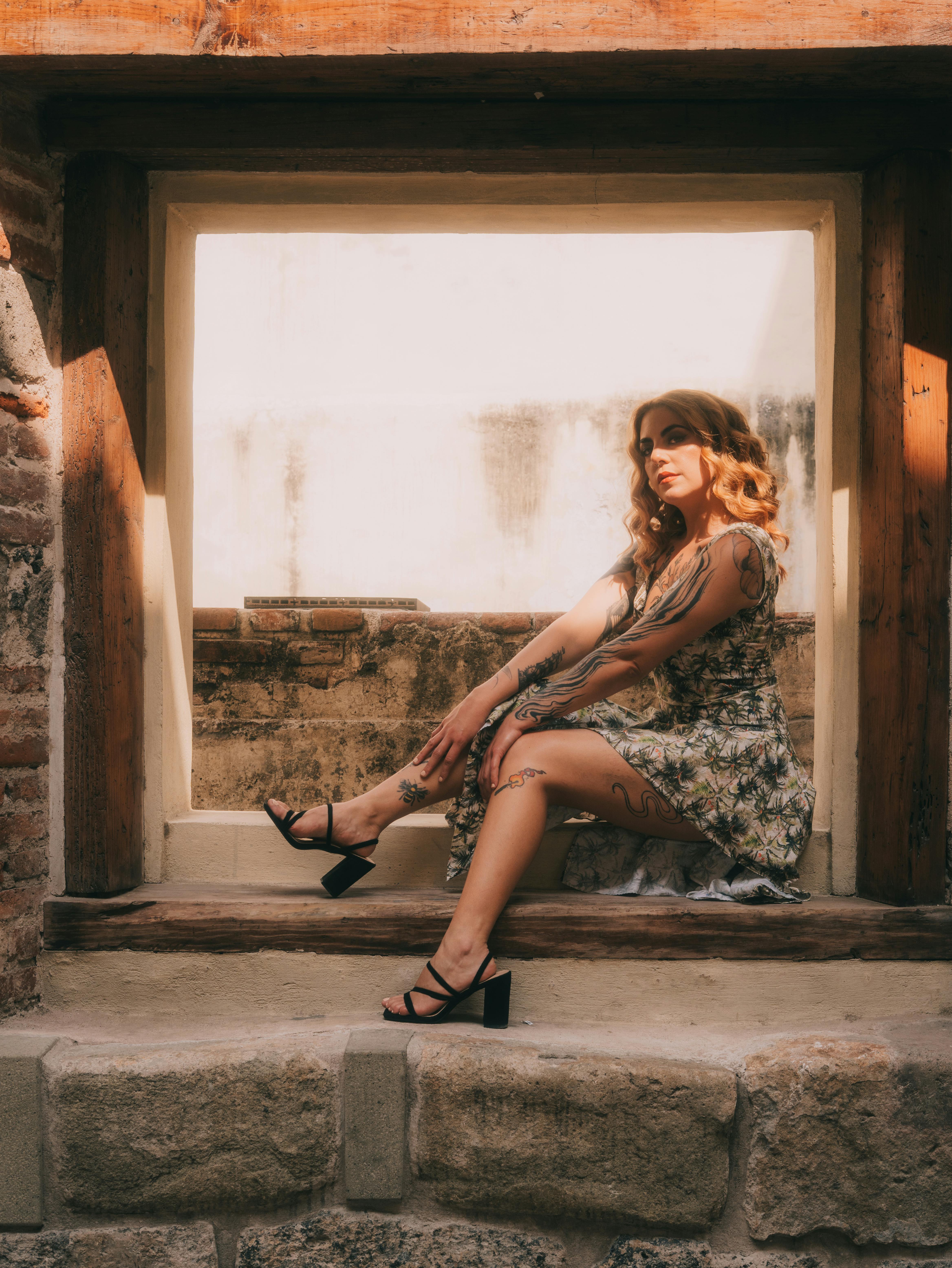 Gratis Una mujer elegante con un vestido floral posa con elegancia en una ventana rústica con suave luz solar. Foto de stock