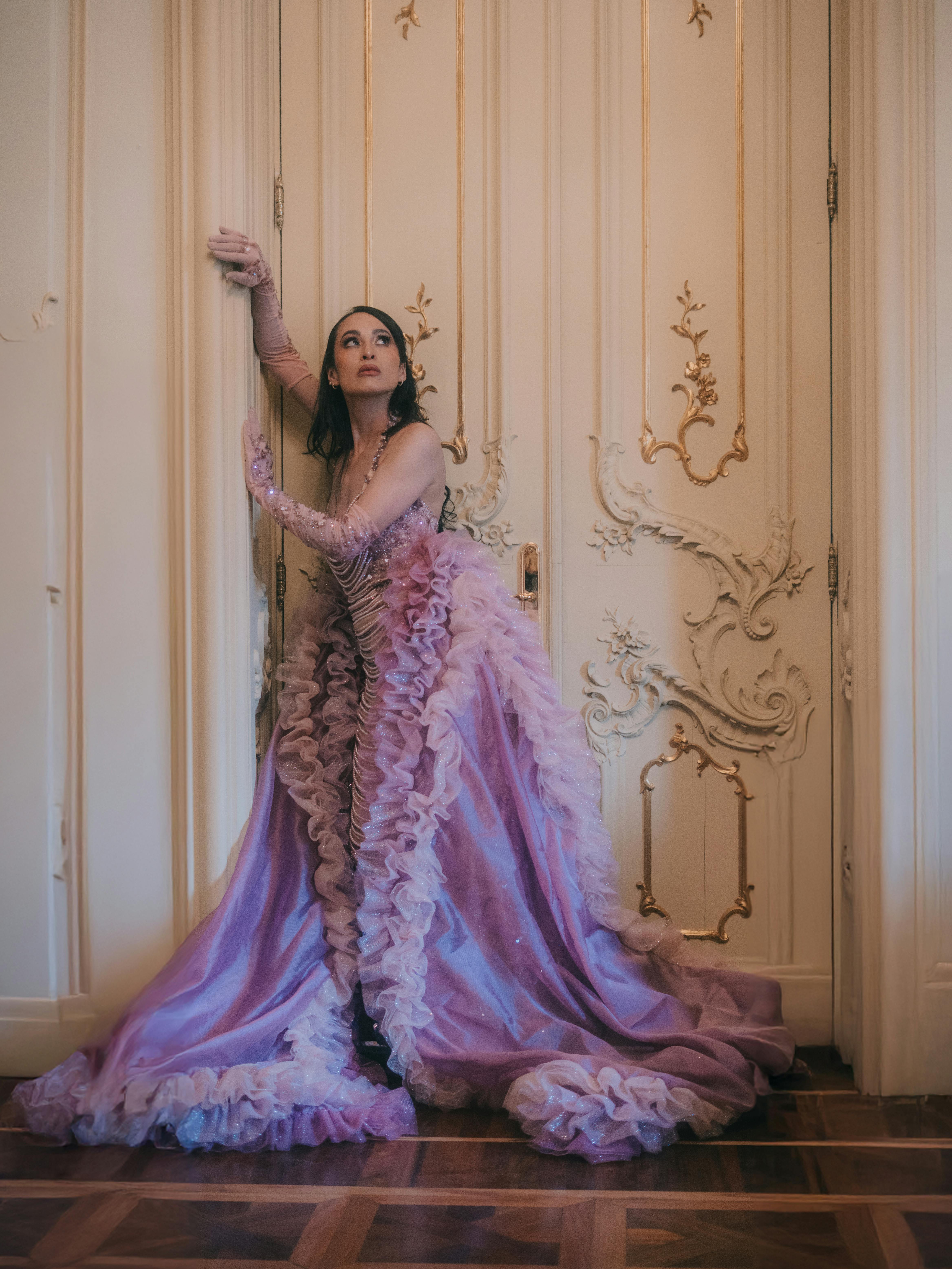 Gratis Una donna con un lussuoso abito viola posa con stile contro una porta decorata all'interno. Foto a disposizione