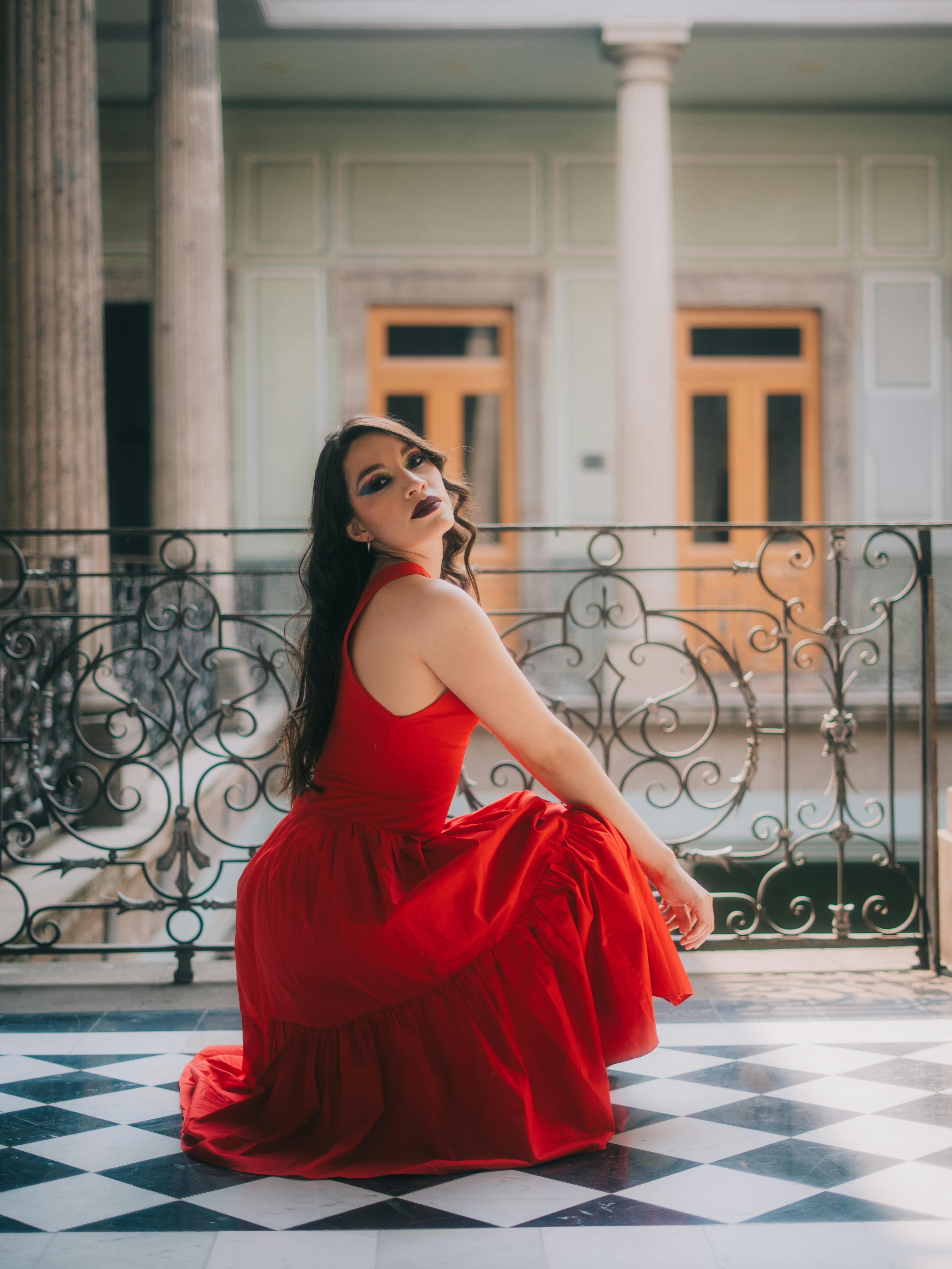 Free Woman in a vibrant red dress posing elegantly indoors with a classic backdrop. Stock Photo