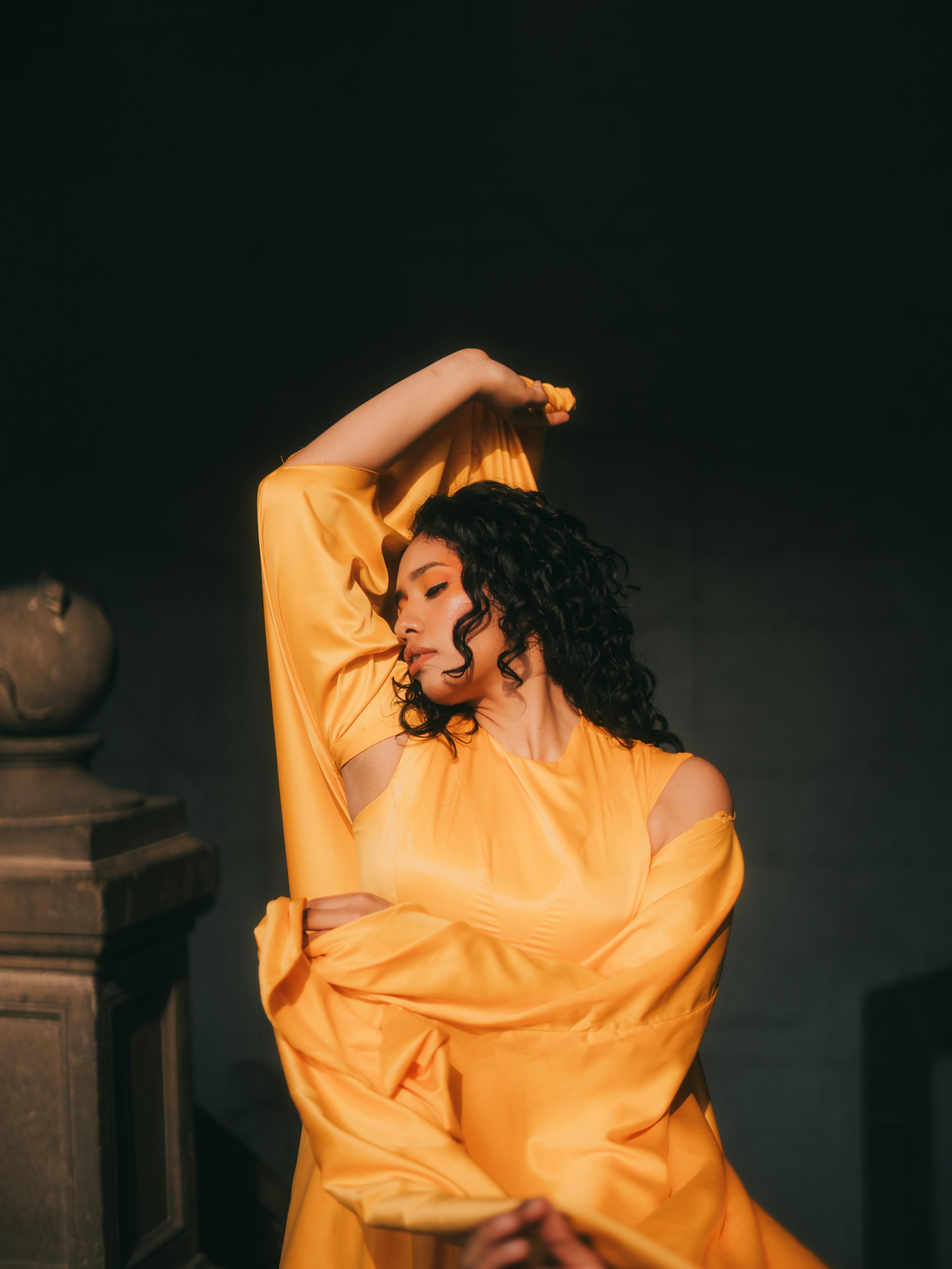 Gratis Seorang wanita dengan gaun kuning mencolok berpose anggun di dalam ruangan, bermandikan cahaya hangat. Foto Stok