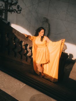 Elegant woman in a flowing yellow dress poses gracefully on a grand staircase with dramatic lighting.