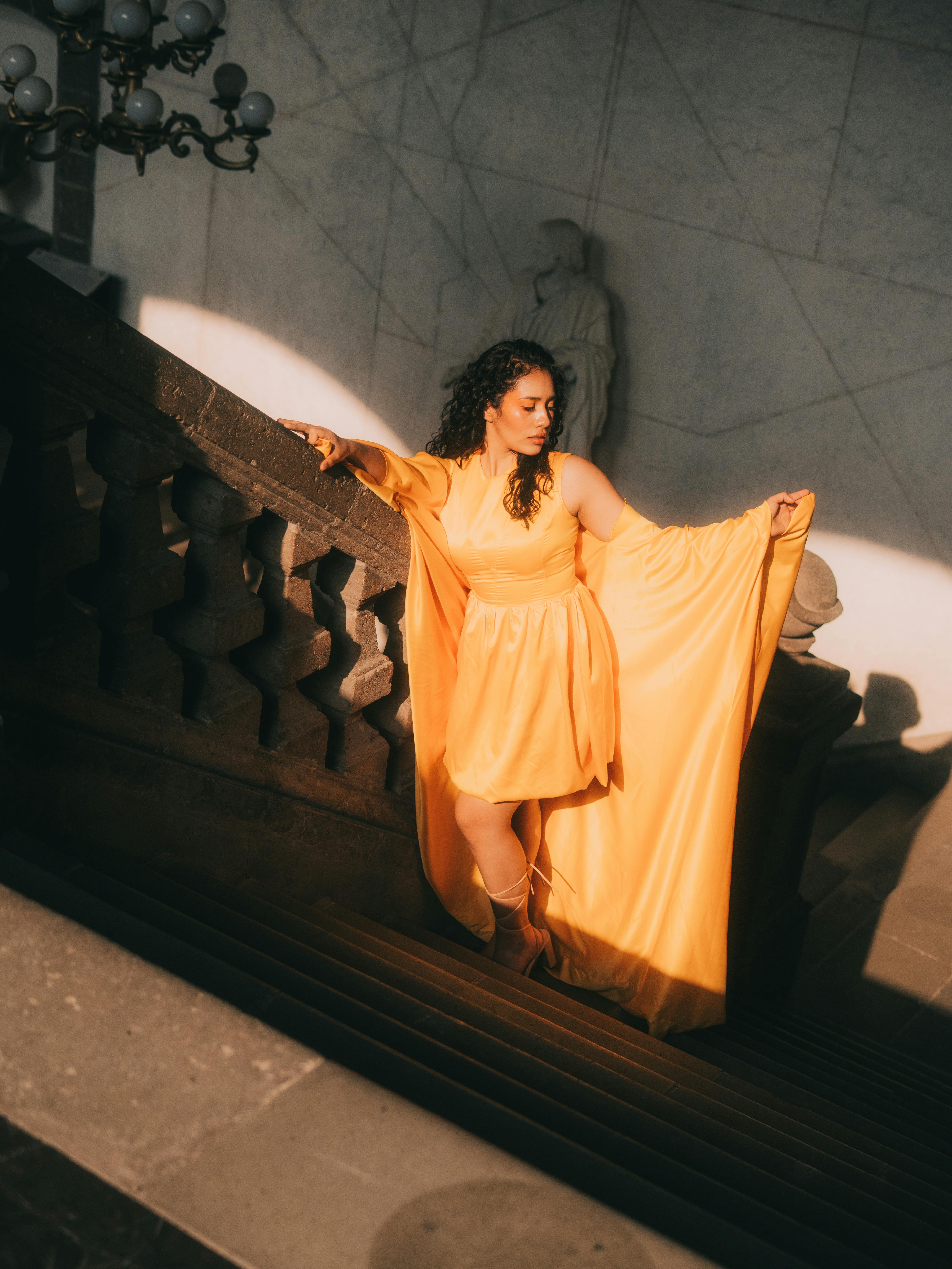 Free Elegant woman in a flowing yellow dress poses gracefully on a grand staircase with dramatic lighting. Stock Photo