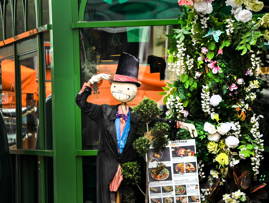 A whimsical display with a mannequin in a top hat, floral decor, and menu at an outdoor restaurant in Krasnodar, Russia.