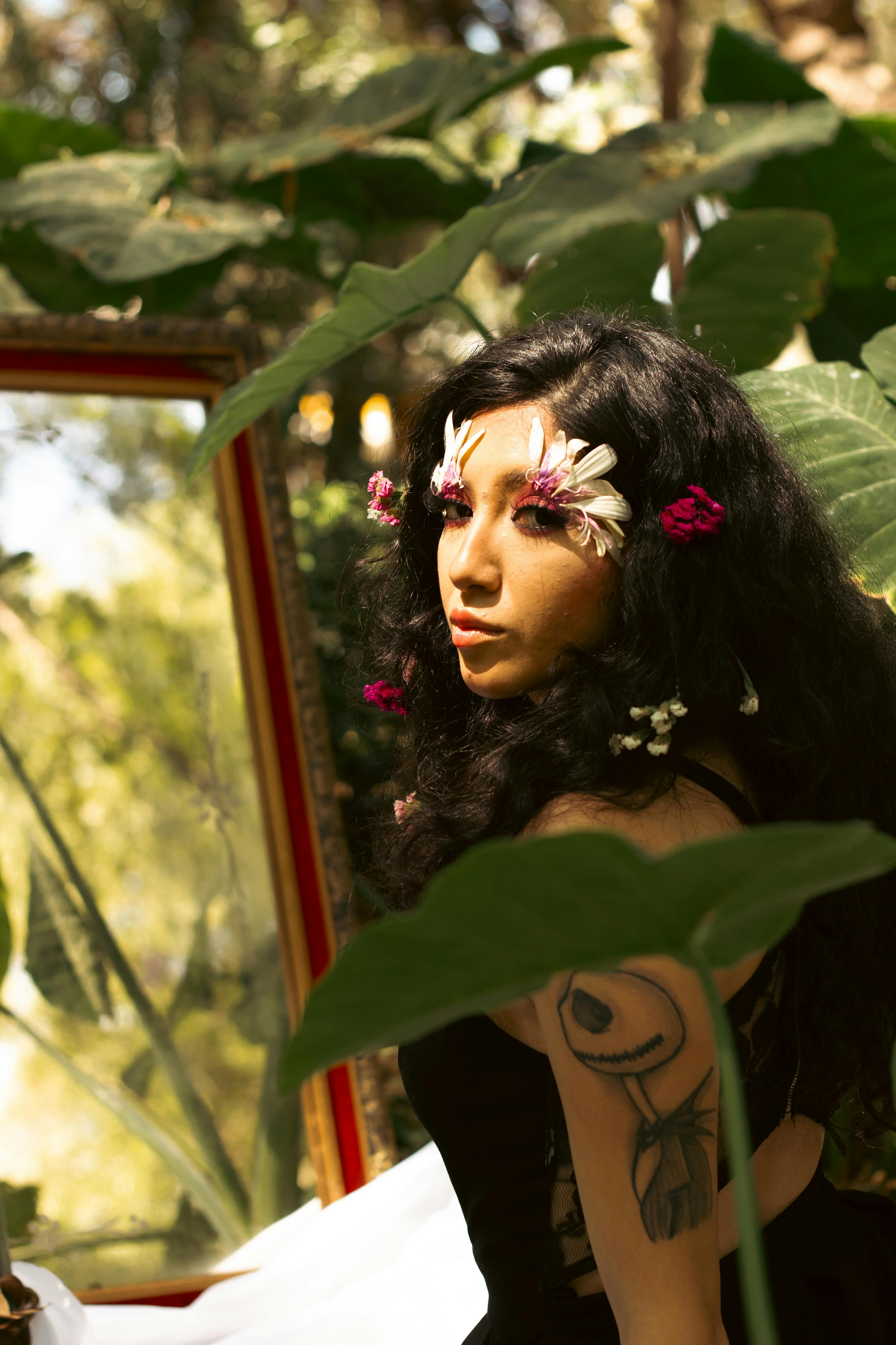 Woman with floral accents poses amidst lush green leaves with sunlight casting soft shadows.
