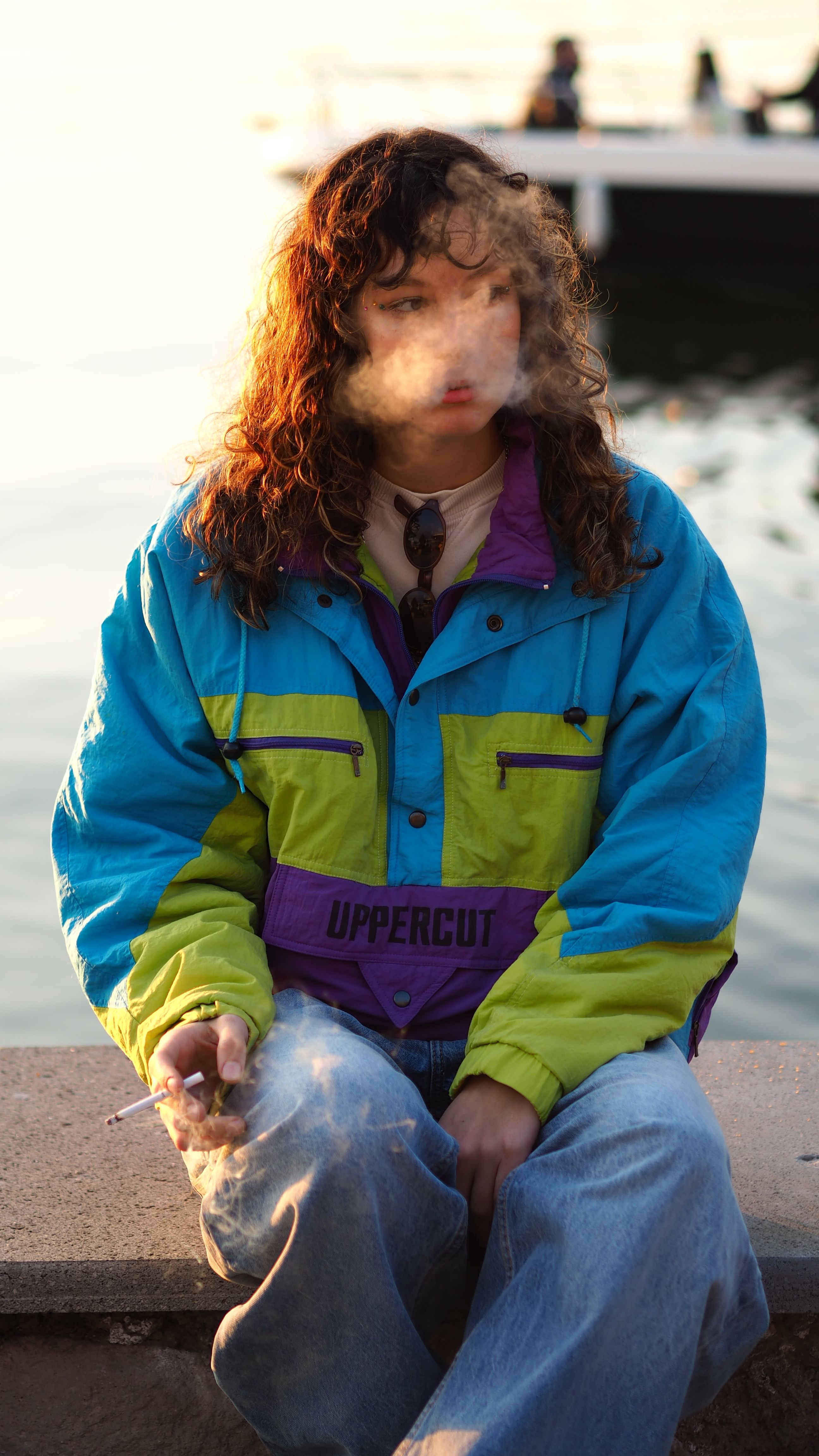 A contemplative young woman smokes by a lake at sunset, wearing a vibrant jacket.
