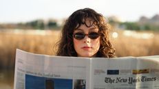 Young woman wearing sunglasses reading a newspaper in a natural outdoor setting.