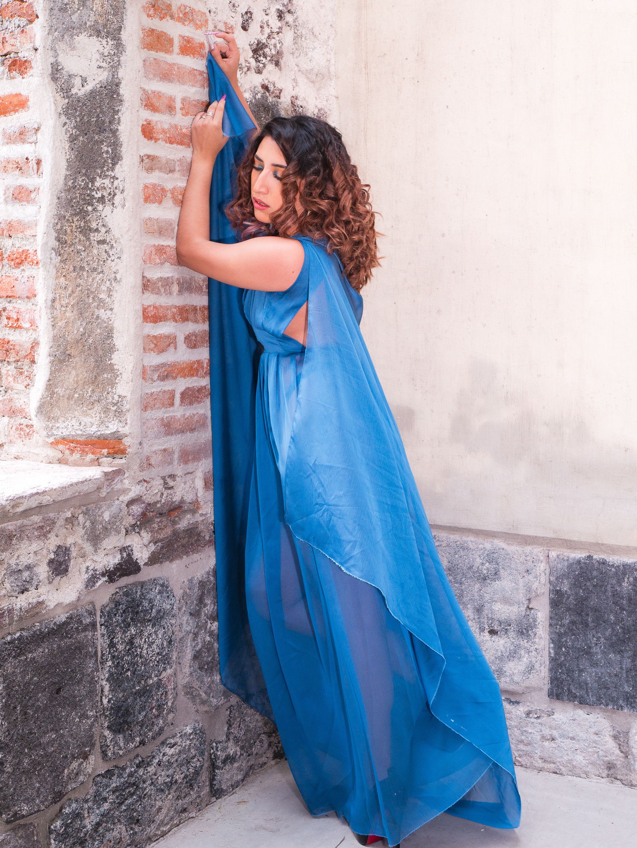 A woman in an elegant blue dress poses against a rustic brick wall.