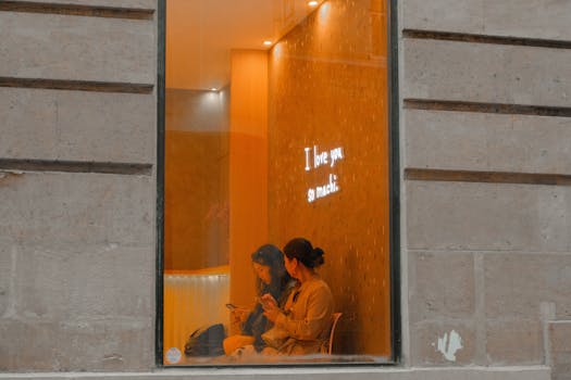 Two women sit in a cozy cafe, viewed through a window with a neon 'I love you so much' sign.