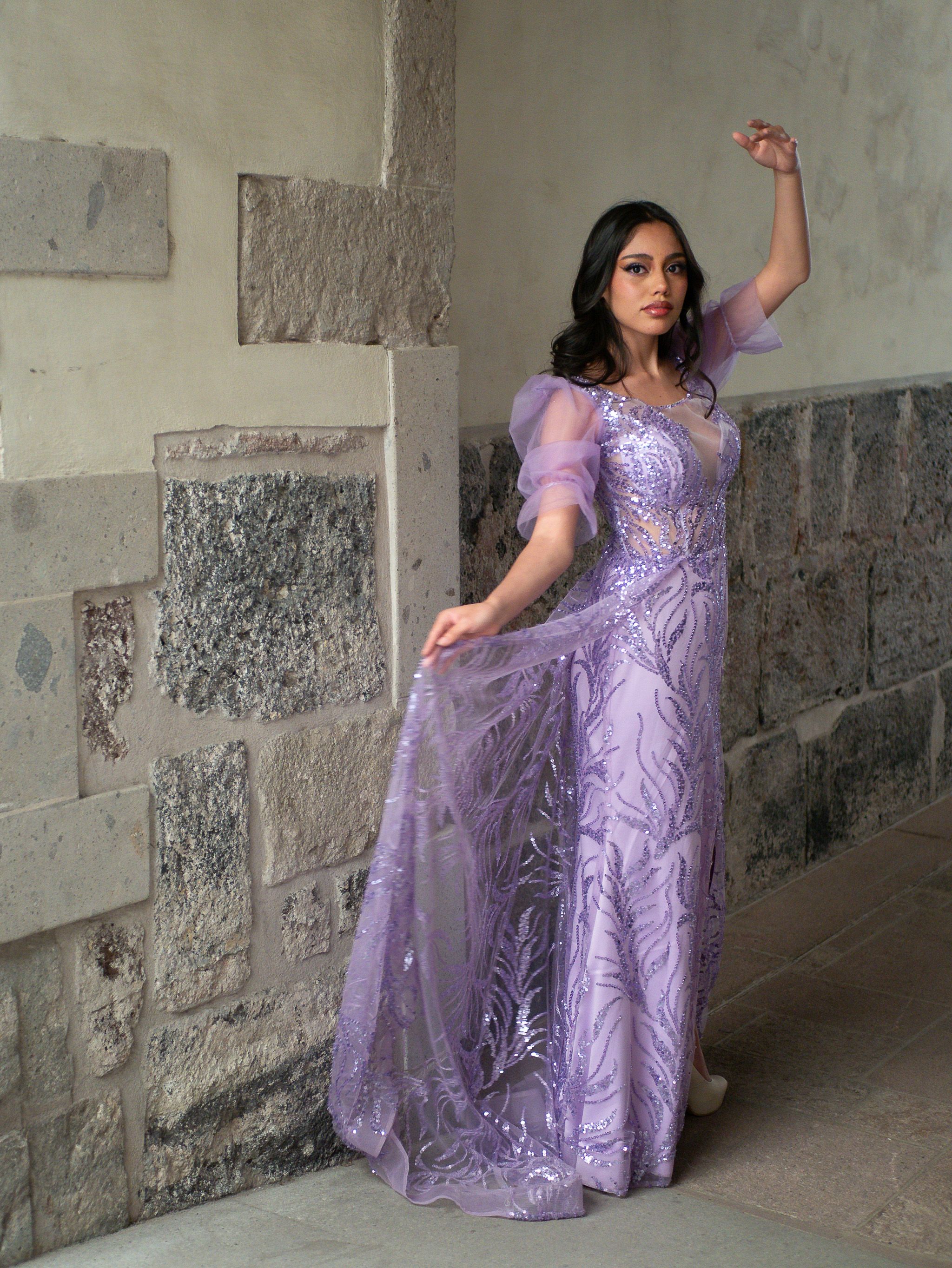 Woman in a lavish lavender gown posing elegantly against a stone wall indoors.