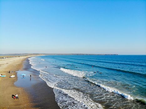 A stunning aerial shot of Tramore Beach in Waterford, Ireland, showcasing surfers and sunbathers under clear blue skies.