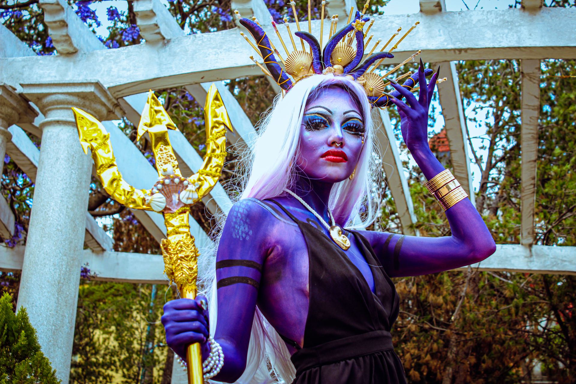 Dramatic purple-skinned cosplay character with ornate headdress and trident in an outdoor setting.